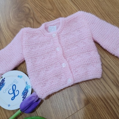 Cardigan rosa bebê em malha com botões brancos sobre chão de madeira, flor roxa e etiqueta redonda branca perto.
