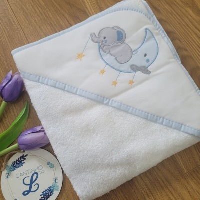 Toalha de bebé branca com capuz bordado com elefante e lua, sobre chão de madeira junto a flores e etiqueta.