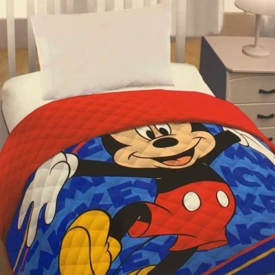 Cama com edredão azul e vermelho com personagem Mickey Mouse num quarto com chão em madeira e móvel branco.