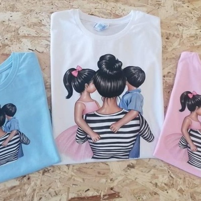 Três t-shirts azul claro, branca e rosa com estampa de mulher com duas crianças vistas de costas sobre fundo de madeira clara
