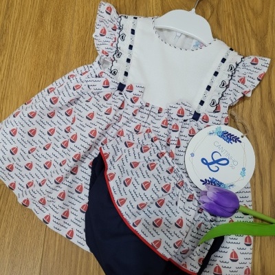 Conjunto infantil com vestido estampado de barcos e calção azul escuro com etiqueta redonda e flor roxa
