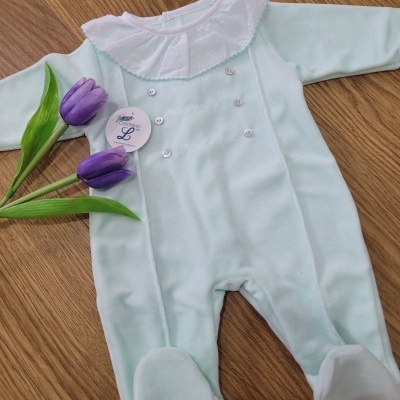 Macacão azul claro de bebé com colarinho branco e botões, ao lado de tulipas roxas em chão de madeira