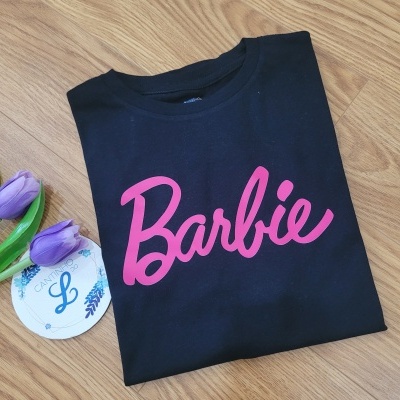 T-shirt preta com texto rosa Barbie ao lado de flores roxas e etiqueta redonda
