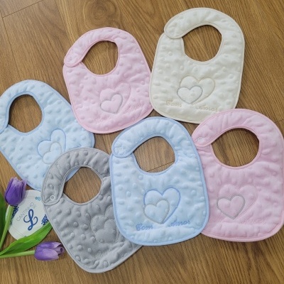 Sete babetes macios de várias cores com coração bordado sobre chão de madeira e duas tulipas