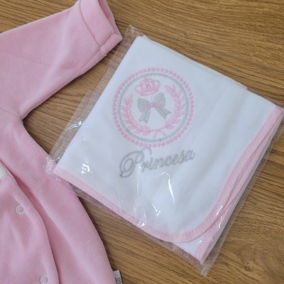 Cobertor branco e rosa com bordado 'Princesa' e peça de roupa cor-de-rosa sobre mesa de madeira