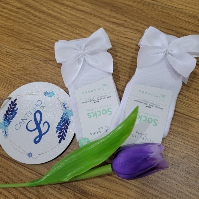 Dois pares de meias brancas de bebé com laços sobre madeira com flor roxa e etiqueta redonda branca
