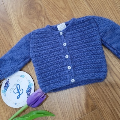 Casaco de malha azul com botões brancos sobre pavimento de madeira com tulipa roxa e embalagem redonda