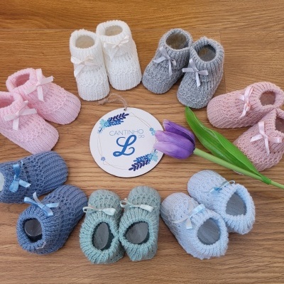 Sapatinhos de bebé coloridos em malha com laços e textura de tricô dispostos em círculo junto a flor lilás e etiqueta CANTINHO L