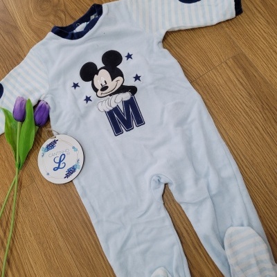 Pijama de bebé azul claro com estampa do Mickey Mouse e detalhes em azul escuro, com tulipas roxas ao lado no chão de madeira.