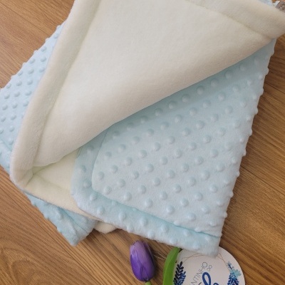 Manta felpuda azul claro com bolinhas de relevo e lado interior branco sobre madeira com flor roxa e etiqueta redonda