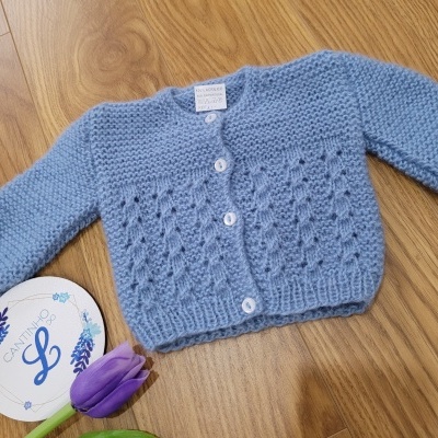 Casaco de malha azul claro para bebé com botões e padrão calado posto sobre madeira com flor roxa e prato branco
