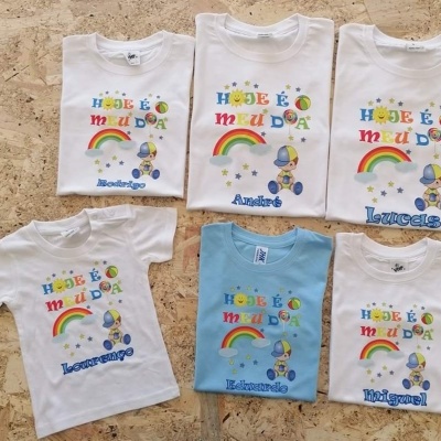 Seis t-shirts infantis com estampado colorido e nomes em azul sobre uma superfície de pedra clara