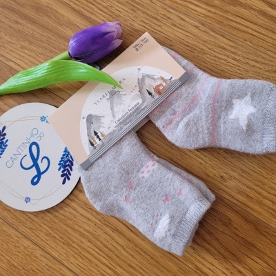 Meias infantis cinzentas com estrelas e corações em fundo de madeira, etiqueta e flor roxa.