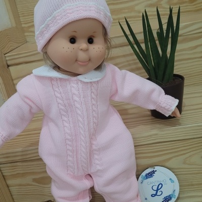 Boneca com roupa de malha cor-de-rosa e gorro, ao lado de vaso com planta e recipiente redondo com texto