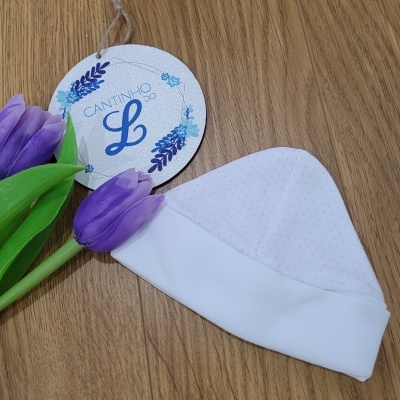Gorro branco de bebé com etiqueta 'CANTINHO L' e duas flores roxas sobre madeira