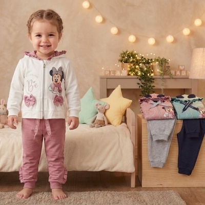 Menina com roupa infantil branca e rosa, ao lado de conjuntos de roupas dobradas com calças cinza e azuis numa decoração de quarto infantil.
