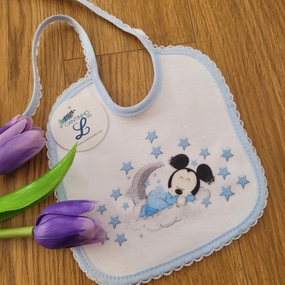 Babete branco e azul claro com imagem de Mickey a dormir e estrelas azuis, com etiqueta redonda, sobre chão de madeira e tulipas roxas.