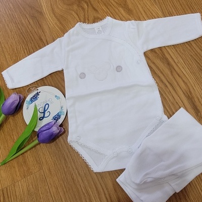 Conjunto de roupa de bebé branco sobre piso de madeira com flores roxas e cartão decorativo