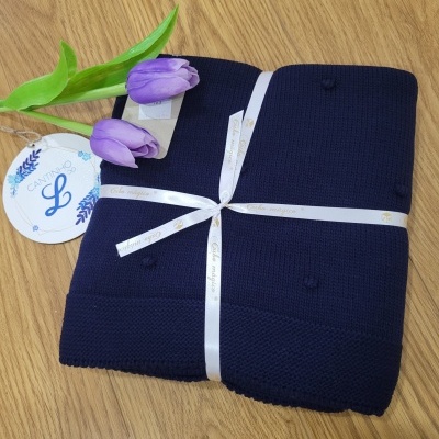 Cobertor azul com pompons, fita branca e etiqueta CANTINHO L sobre superfície de madeira