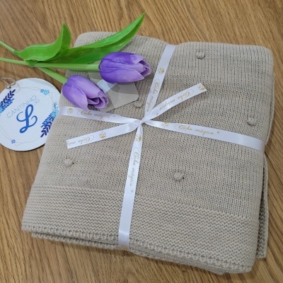 Cobertor bege claro dobrado com fita branca, flores roxas artificiais e etiqueta redonda em superfície de madeira