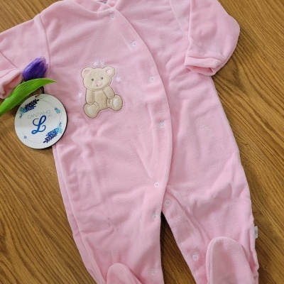 Macacão de bebé rosa com ursinho bordado e botões brancos sobre mesa de madeira.