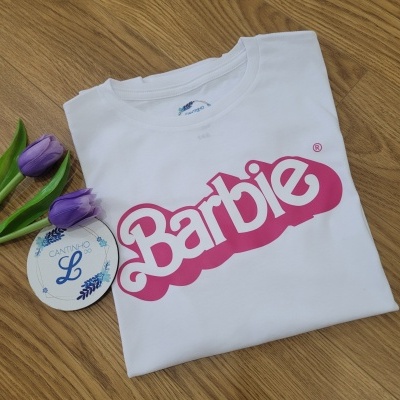 T-shirt branca com logótipo Barbie rosa dobrada sobre superfície de madeira com flores tulipa roxa ao lado