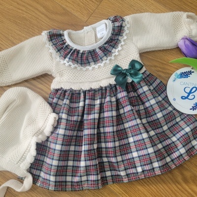 Vestido de bebé com saia xadrez, laço verde e gorro creme sobre chão de madeira