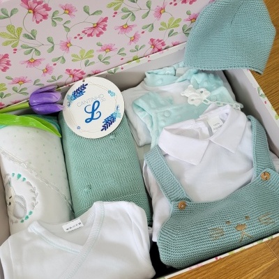 Roupas de bebé em caixa decorada com padrão floral, incluindo colete de tricô verde-água, camisa branca e gorro.