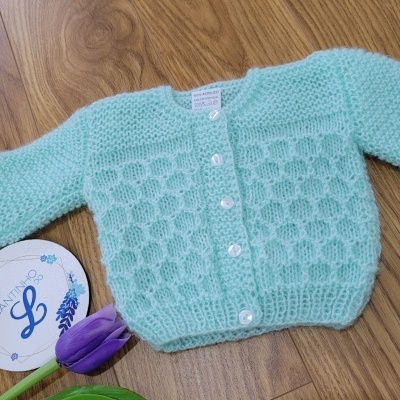 Cardigan de bebé azul claro com botões brancos sobre madeira, cartão redondo e flor roxa