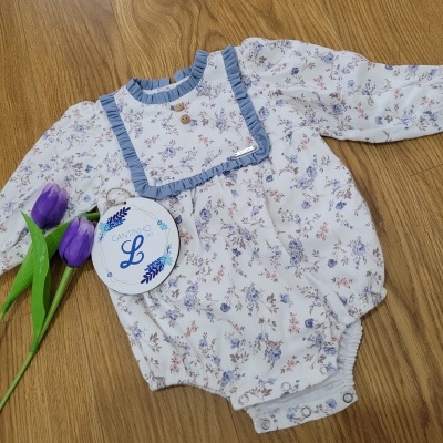 Body de bebé floral branco com folho azul e botões de madeira em superfície de madeira com tulipas roxas