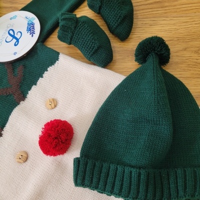 Conjunto de roupa de bebé em malha verde e bege com pompom vermelho, botões e acessórios em cima de superfície de madeira.