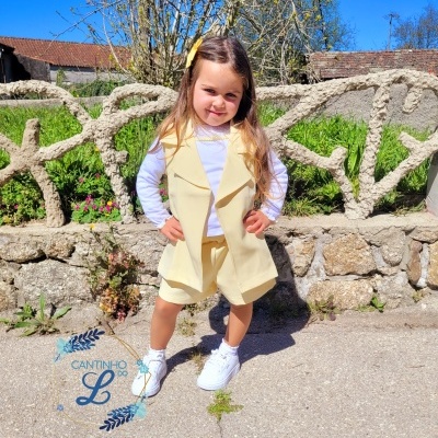 Menina com roupa bege e branca posando em ambiente exterior com muralha de pedra