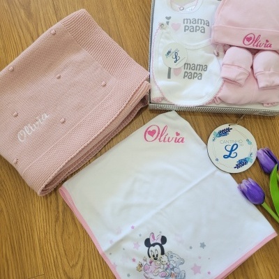 Conjunto de bebé com manta tricotada rosa, babete, gorro e peúgas rosa, manta branca com borda rosa e imagem de Minnie, sobre chão de madeira.