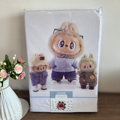 Embalagem de roupa de cama com imagem de três bonecos de pelúcia, vaso branco com flores, superfície de madeira