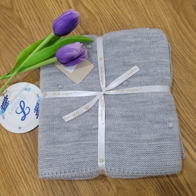 Manta cinzenta de malha com fita 'Carlo Magno', cartão branco 'L' e flores roxas sobre madeira.