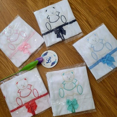 Cinco fraldas brancas bordadas com ursinhos coloridos e laços sobre chão de madeira, com uma flor roxa e um cartão