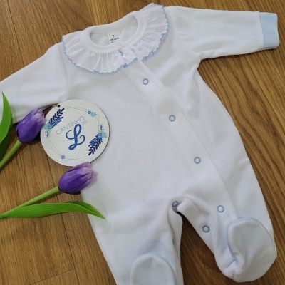 Macacão branco para bebé com golinha e botões azuis sobre superfície de madeira com flores roxas e círculo decorativo