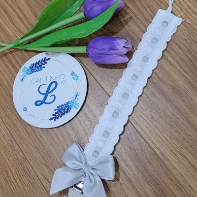 Laço branco com presilha ursinho em madeira, flores roxas artificiais e cartão CANTINHO DO L sobre superfície de madeira