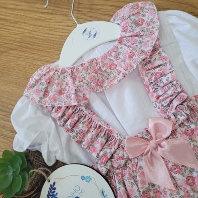 Vestido infantil floral rosa e branco com babados e laço cor de rosa num cabide branco sobre madeira