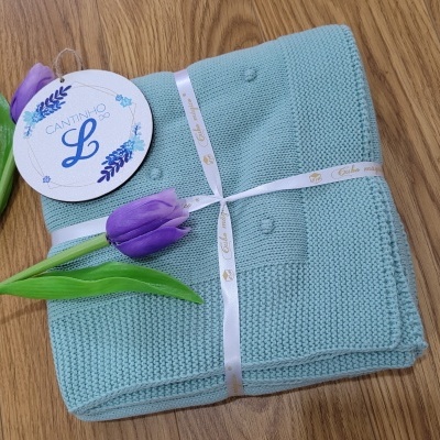 Manta azul clara com pompons, etiqueta 'CANTINHO L', fita branca dourada e flores roxas sobre chão de madeira