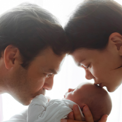 Homem e mulher beijam um bebé vestido de branco em fundo claro