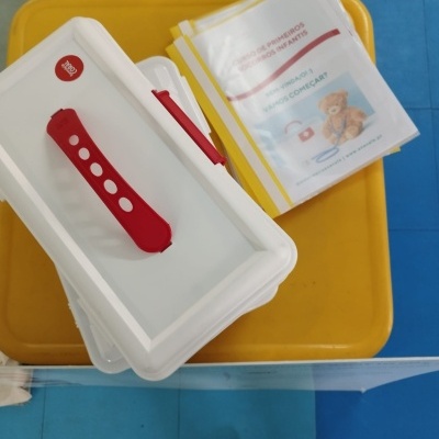 Caixa plástica branca com tampa vermelha e folhas informativas com ursinho em superfície amarela.