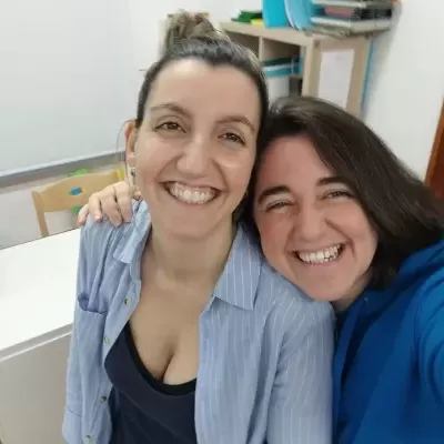 Duas mulheres sorridentes tiram selfie em interior com mesa e prateleiras ao fundo