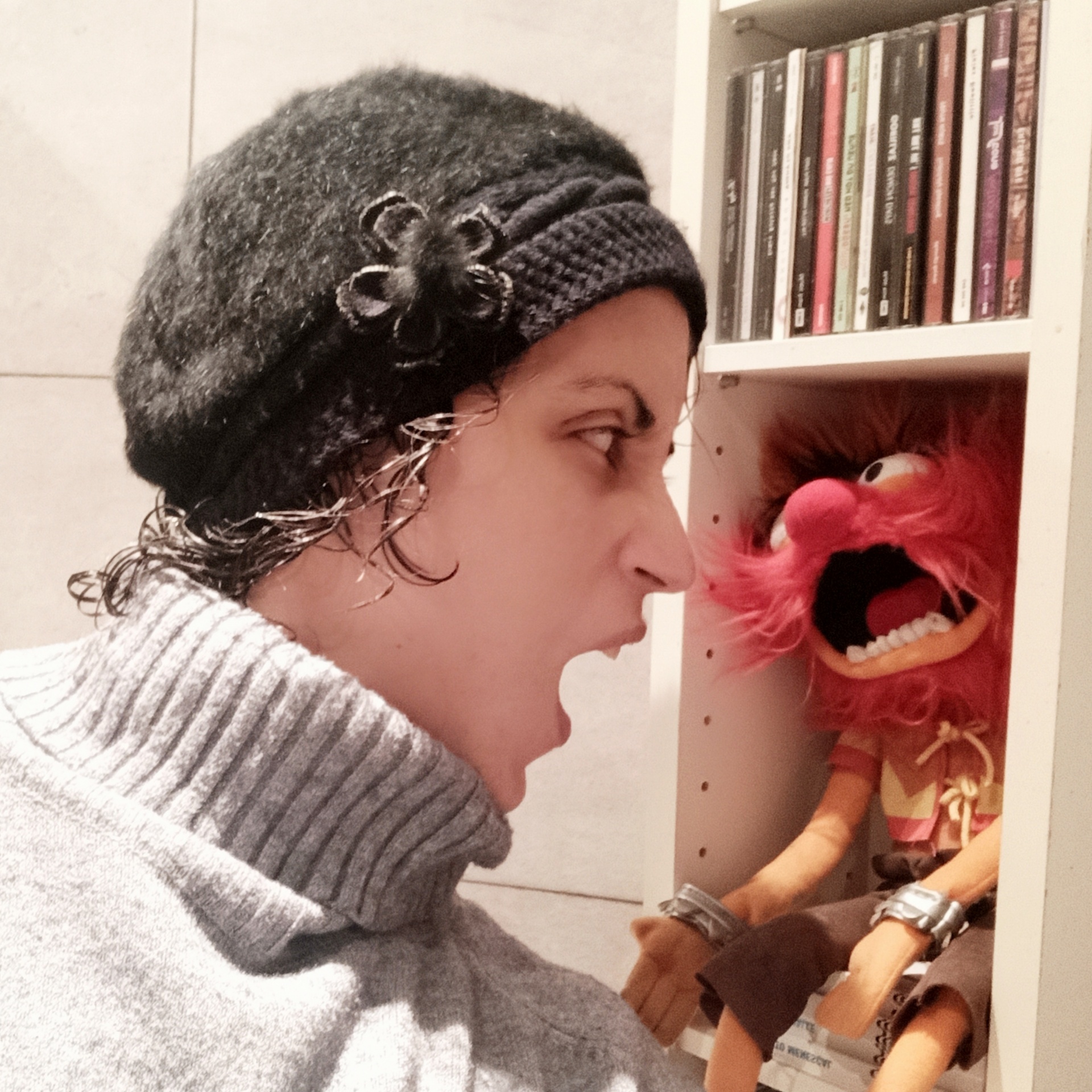 Mulher usando gorro preto e suéter cinza, olhando para marioneta vermelha em estante branca com CDs