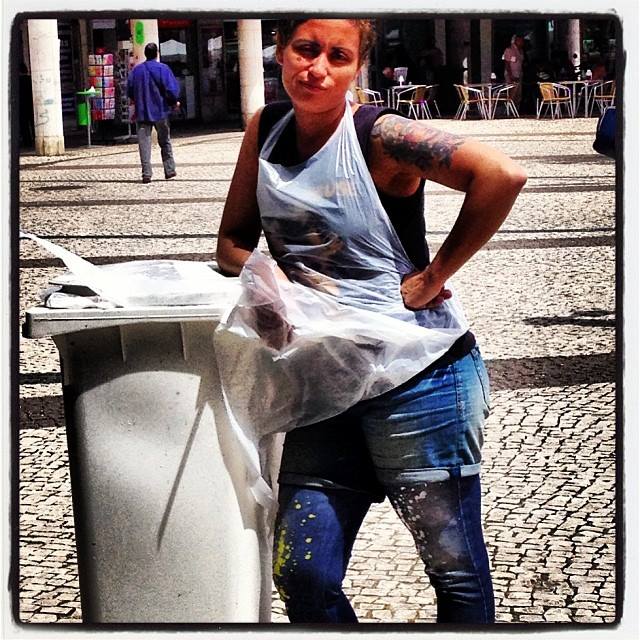 JUNTAR DINHEIRO ... URGENTE Mulher com avental plástico e calças de ganga com manchas junto a contentor de lixo em calçada portuguesa