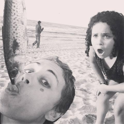 Duas pessoas numa praia a interagir com um peixe, com uma terceira pessoa ao fundo perto da água.