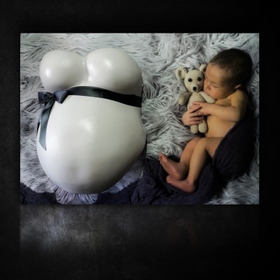 Busto de gesso branco com fita preta ao lado de bebé a segurar ursinho sobre cobertor cinzento