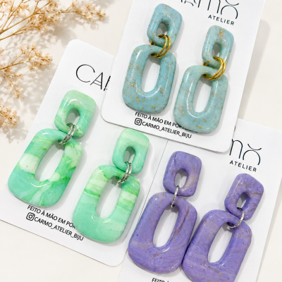 Três pares de brincos pendentes em resina colorida, embalados em cartões brancos com texto da marca, sobre superfície clara com flores secas ao lado.