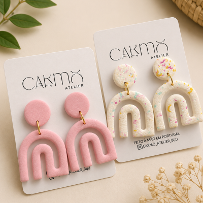 Dois pares de brincos de arco, um rosa e outro branco com manchas, em cartões CAKMO ATELIER sobre fundo claro com folhas e flores secas.