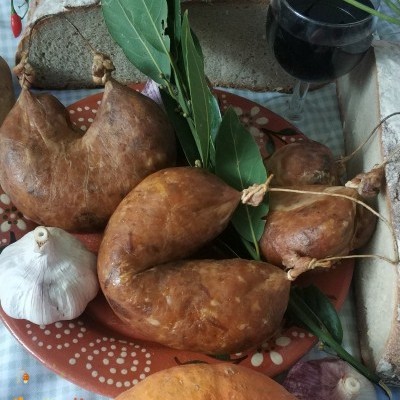 Prato com enchidos, alho, folhas verdes, pão, copo de vinho tinto e abóbora sobre pano xadrez.